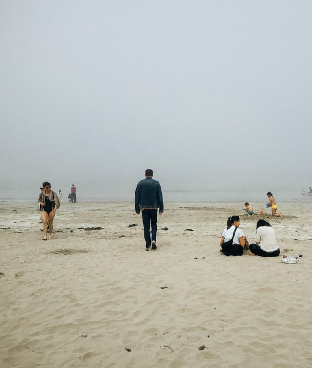 A fully-clothed man walks away from the camera across a sandy beach towards the sea. Other children are playing, unaware of him.