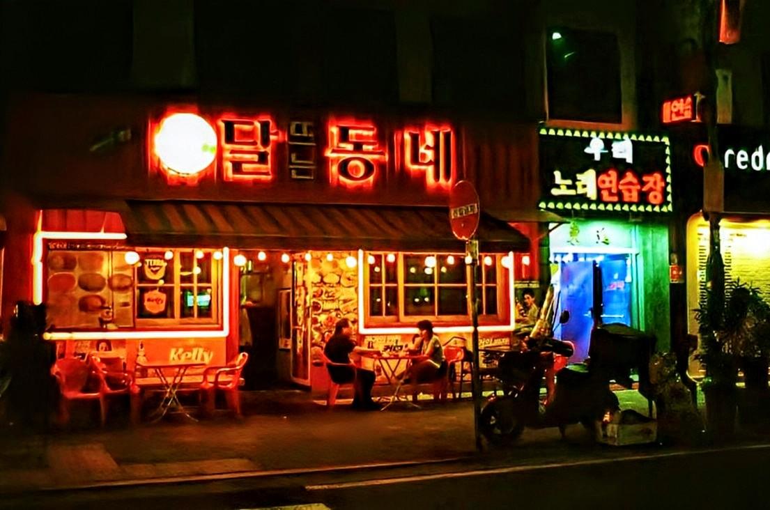Colour 35mm film photograph of a couple enjoying a meal outside a brightly lit restaurant. The shops next door are also brightly lit. Taken in Seoul, South Korea, June 2025.
