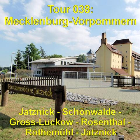 Gelb gestrichenes, mehrstöckiges Gebäude mit Satteldach und seitlichem Turmteil. Daneben steht ein flacher, moderner Anbau mit großen Fenstern. Das Gelände ist von einem Metallzaun mit geschlossenem Tor umgeben. Im Vordergrund befindet sich ein Holzzaun mit einem breiten Holzschild. Ein gepflasterter Weg führt zum Eingang. Rasenflächen und einige Bäume rahmen die Anlage ein. Der Himmel ist blau mit lockeren weißen Wolken. Die Gebäude wirken gewerblich oder industriell genutzt.