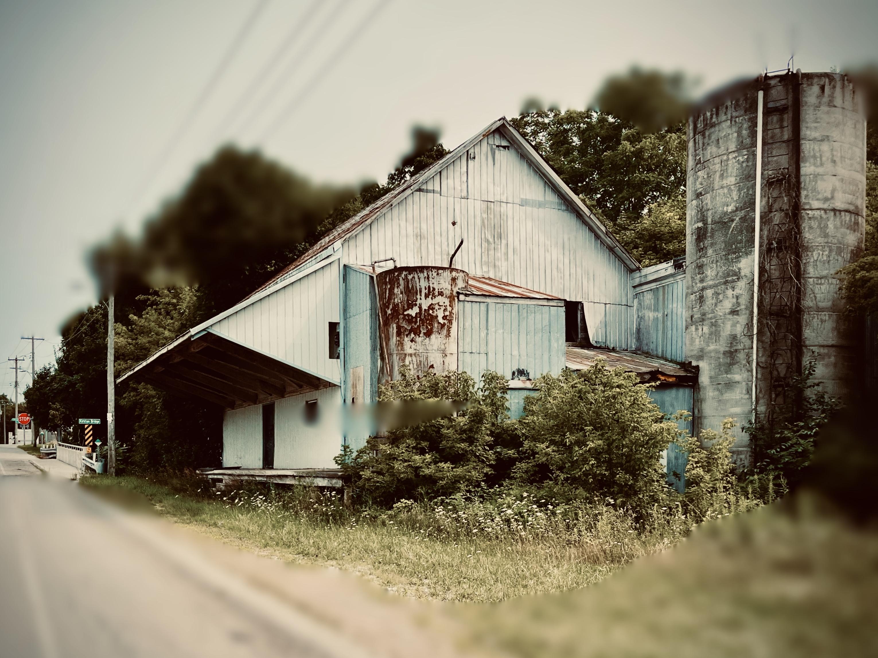 A building probably constructed in the 1940's or 50's when the highway system was in development, now dilapidated, abandoned and locked. Weeds and rubble line its foundation, its metal roof is rusting and decades old formerly white paint is streaked with stains. An overhang casts a long shadow over a concrete riser running its full frontal width and behind some bushes nature has installed, it appears connected to an ancient concrete silo that's about a third of the height of any serious farm silo. One can only guess at its former ownership and purpose.