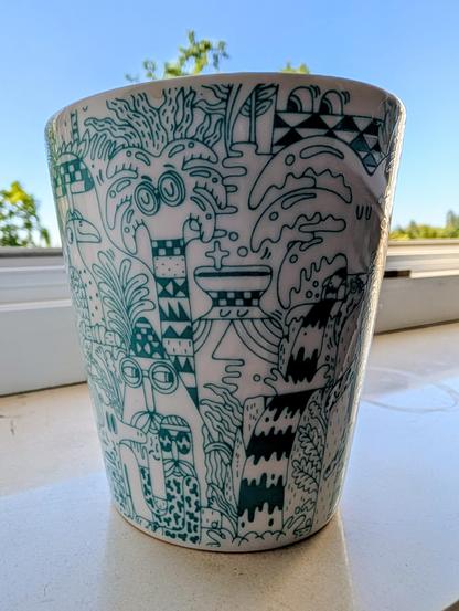 Weißer Keramik-Übertopf auf einer Fensterbank, bedruckt mit detaillierten, dunkelgrünen Doodling-Illustrationen im Comic- und Dschungel-Stil. Zu sehen sind stilisierte Palmen mit Gesichtern, Fantasiewesen, Pflanzen und geometrische Muster. Im Hintergrund ist ein Fensterrahmen mit Blick auf einen blauen Himmel und grüne Baumkronen zu erkennen.