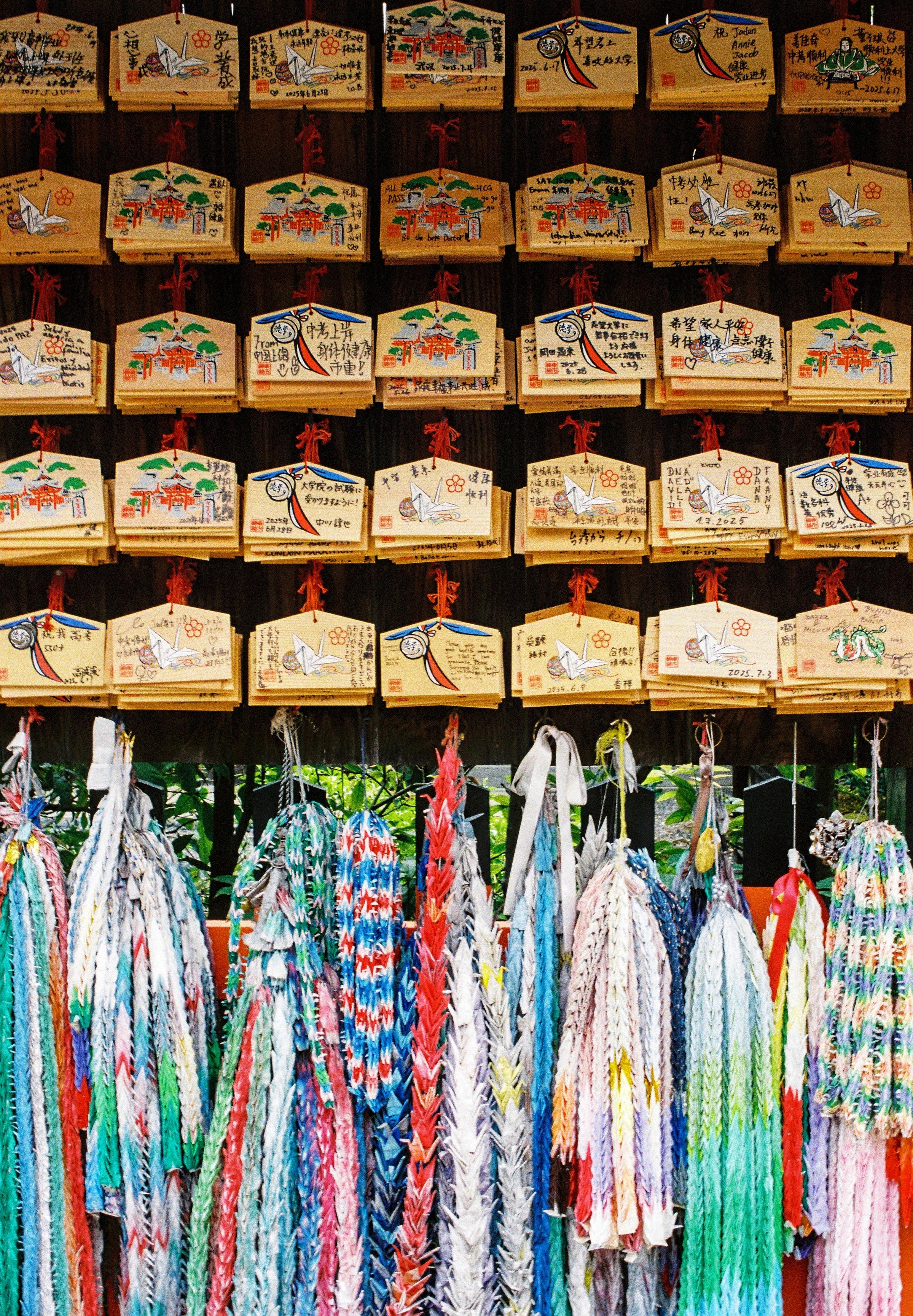 Colour 35mm film photograph of a display in a souvenir shop in Kyoto, Japan.
Image shows wooden plaques which you could buy and add a message to. Underneath are colourful played cloths.
Behind you can just see trees.