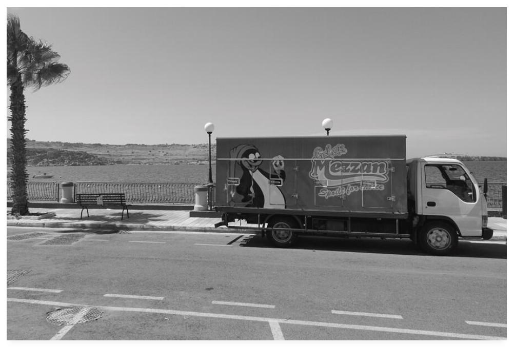 Black and white photograph of a refrigerated truck parked up at the coast.  On the side is a picture of a cartoon penguin holding a cornet.