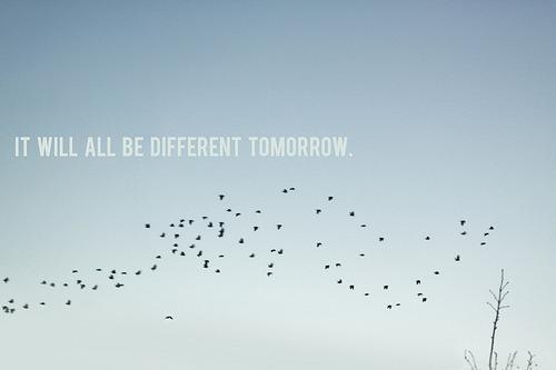 A scattered flock of dark birds flying across a pale blue sky. White, capitalized text overlaid on the image reads, 'IT WILL ALL BE DIFFERENT TOMORROW.' A few bare tree branches reach up into the frame from the bottom right corner.