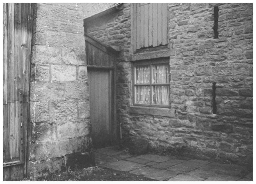 Vintage looking black and white film photograph showing the rear of a farmhouse with a wooden door alongside a four pane window.