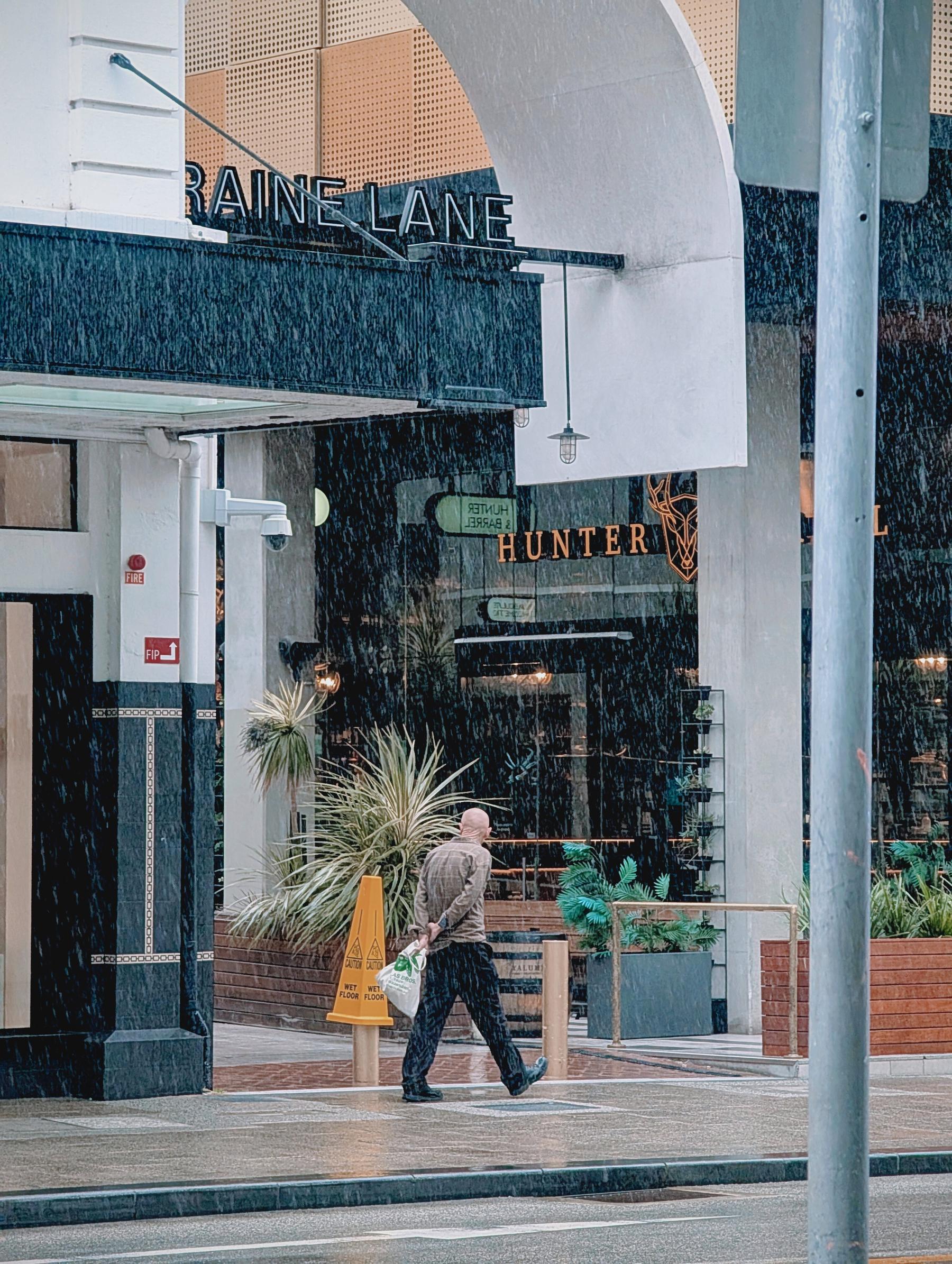Auto-generated description: A person holding a bag is walking on a wet sidewalk in front of a building with a HUNTER sign during heavy rain.