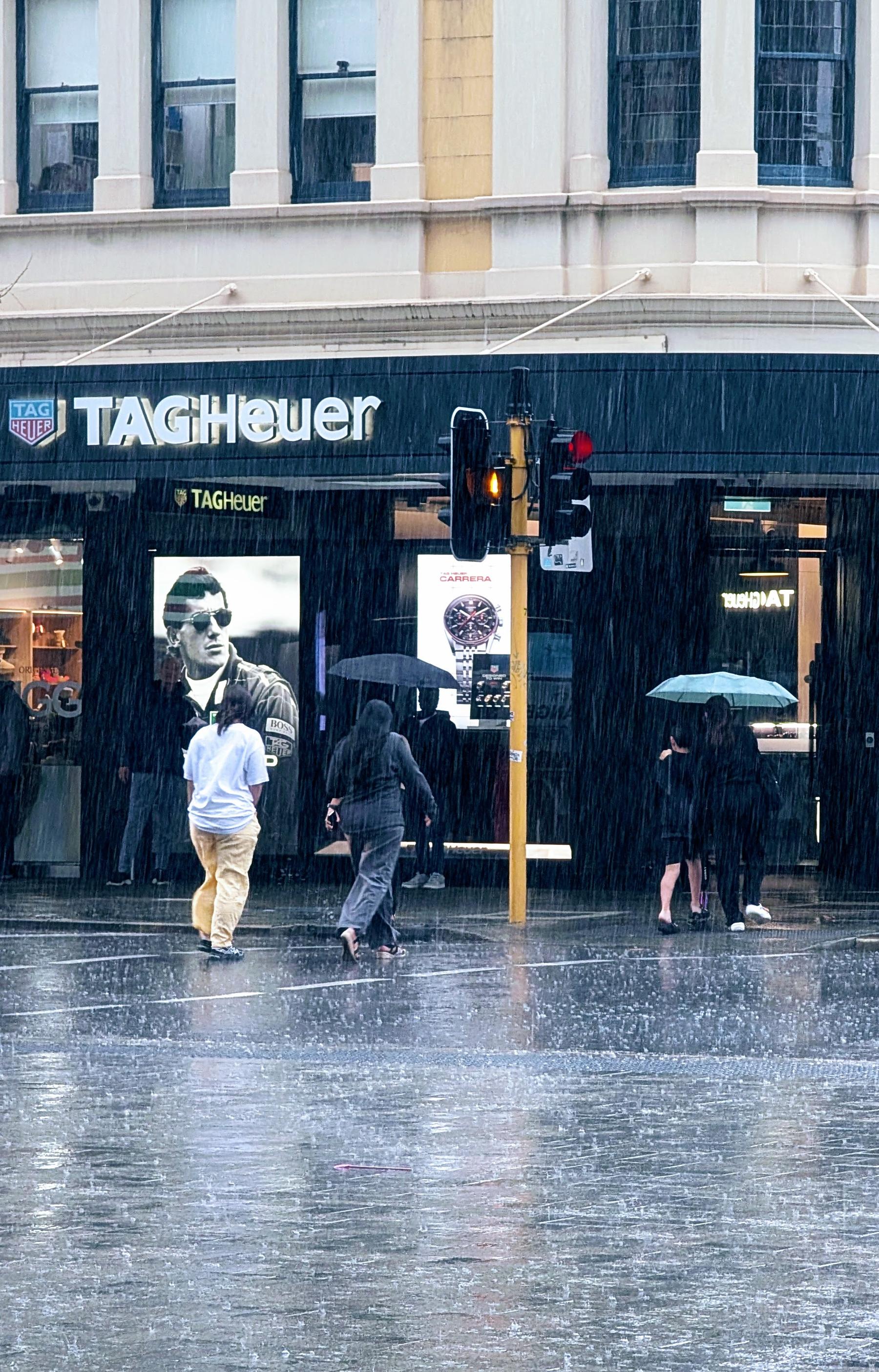 Auto-generated description: People are crossing a rainy street in front of a TAG Heuer store, some holding umbrellas while others do not.