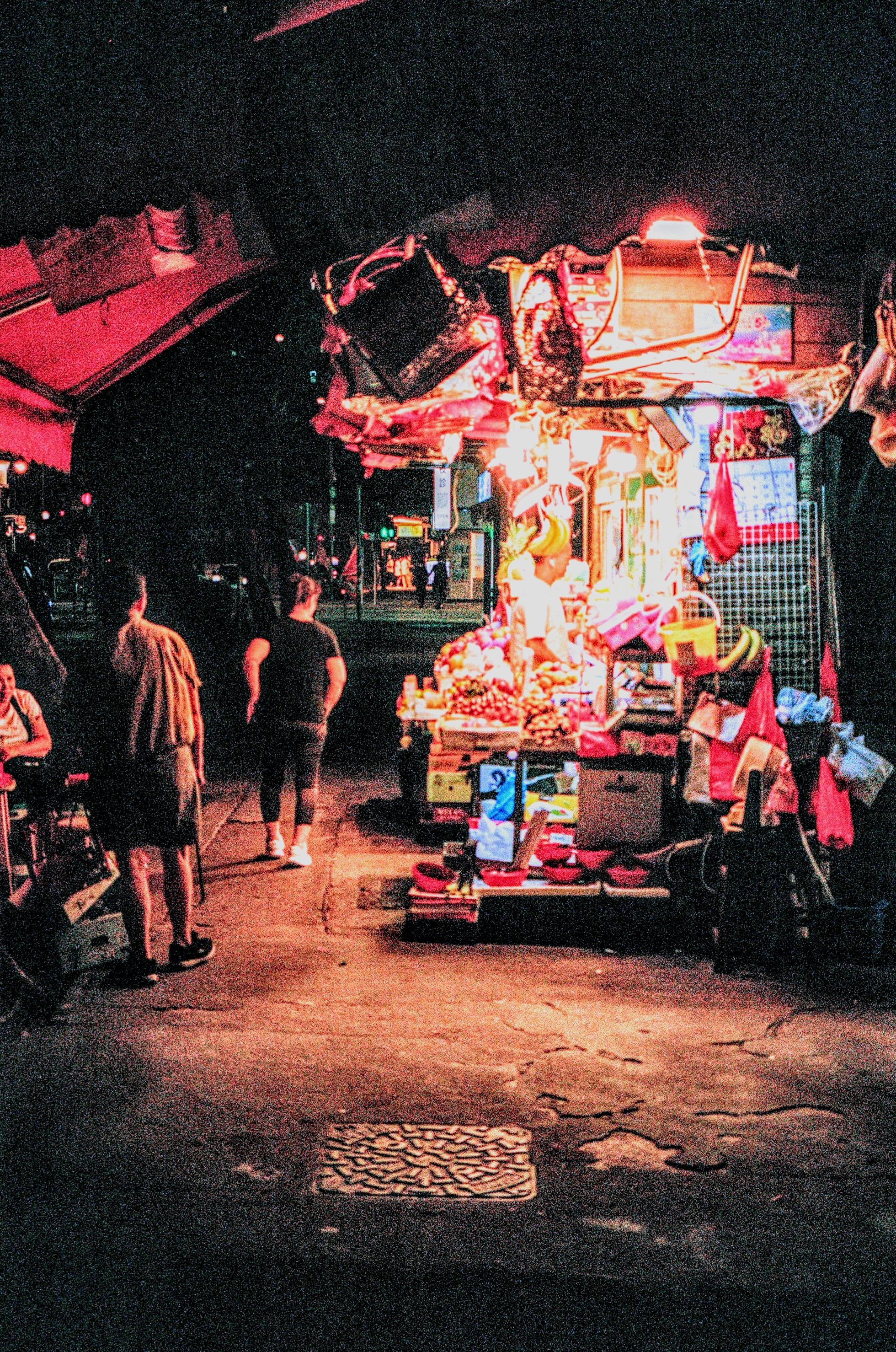 Colour 35mm film photograph of a fruit and vegetable market stall in Hong Kong during the evening. Photo shows a person buying goods at the brightly lit stall as three other people look on on the sidewalk.