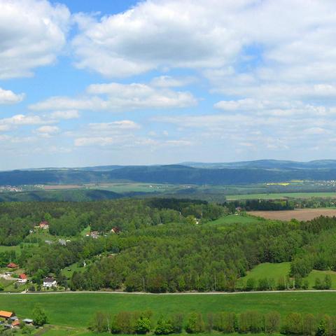Eine weite Landschaft mit sanften Hügeln, Wiesen, Feldern und verstreuten Wäldern erstreckt sich unter einem teils bewölkten Himmel. In der unteren Bildhälfte sind einzelne Häuser und Gehöfte zwischen grünen Flächen und kleinen Waldstücken zu sehen. Im Hintergrund verlaufen langgestreckte, bewaldete Höhenzüge, die sich bis zum Horizont ziehen. Der Himmel ist von zahlreichen weißen, bauschigen Wolken durchzogen, die Schatten auf die Landschaft werfen.