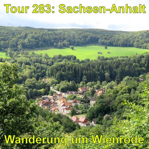 Blick von einem Aussichtspunkt auf ein Dorf in einem Tal, umgeben von dicht bewaldeten Hügeln. Die Dächer der Häuser sind vorwiegend rot, und die Gebäude gruppieren sich entlang einer Straße. Jenseits des Dorfes öffnet sich eine große Wiese auf den Hügeln. Das Grün der Bäume und Wiesen ist leuchtend und vielfältig. Das Licht ist klar.