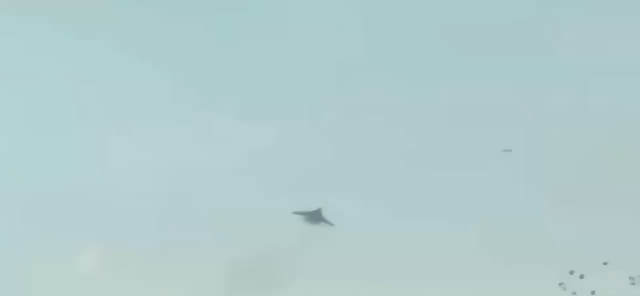 A fighter plane pursues a triangle shaped drone in flight, the fighter banks in front of the drone, making the air unstable, the drone loses enough lift and points down and impacts the ground. Orange flames billow up from the drone’s impact. The fighter turns and flies passed our view as they regroup. 