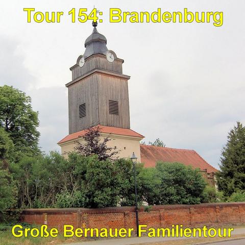 Ein rechteckiger Kirchturm mit vertikaler Holzverkleidung erhebt sich über einem niedrigen, hell verputzten Gebäude mit rotem Ziegeldach. Am oberen Teil des Turms sind auf mehreren Seiten runde Zifferblätter angebracht. Darüber sitzt eine geschwungene Turmhaube mit kleiner Spitze und Wetterfahne. Schmale Lüftungsöffnungen sind in die Holzfassade eingelassen. Das Gebäude ist von dichtem Grün umgeben, darunter Büsche und hohe Bäume. Im Vordergrund verläuft eine niedrige Backsteinmauer entlang ein…