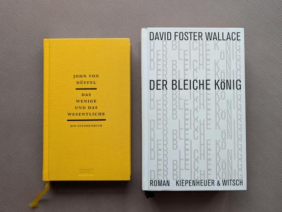 Zwei Bücher liegen nebeneinander auf einem grauen, fein strukturierten Untergrund. Das linke Buch hat einen senfgelben Stoffeinband mit schwarzer Aufschrift: Oben steht "JOHN VON DÜFFEL", in der Mitte der Titel "DAS WENIGE UND DAS WESENTLICHE", eingefasst von zwei horizontalen Linien, und darunter der Zusatz "EIN STUNDENBUCH". Am unteren Rand ist der Verlagsname "DUMONT" leicht eingeprägt. Ein gelbes Lesezeichenband schaut unten aus den Seiten heraus. Das rechte Buch hat ein weißes Cover mit de…