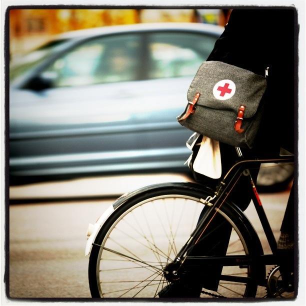 Square image. Top half is mostly a side-profile of a blurry silver-blue car driving right-to-left in the background. Bottom half is the rear wheel of a black-and-white bicycle moving left-to-right, upon which sits a person wearing a black coat, black pants, and black shoes and with a gray messenger bag hanging from their back. The bag has light-brown leather closure straps and a logo of a red cross in a white circle.