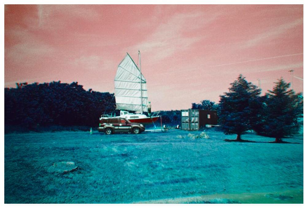 Colour adjusted film photograph showing a chap on the deck of his yacht adjusting a sail.  The yacht is moored in a field beside a parked SUV.  The sky is pink and the grass a bluey green.