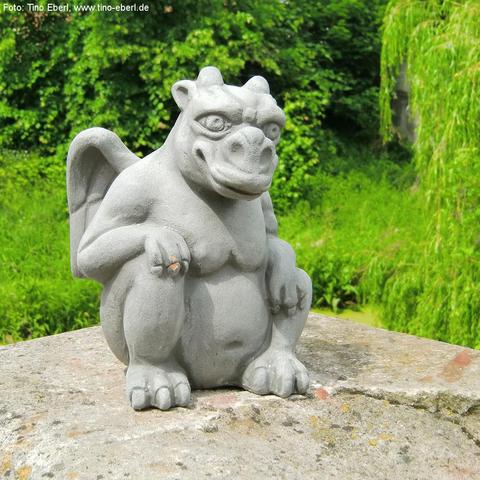 Graue Figur eines geflügelten Fabelwesens mit tierischen Zügen, sitzend auf einem steinernen Sockel. Die Figur hat ein breites Gesicht mit großen Augen, kleinen Hörnern, kräftigen Armen und Klauenhänden. Der Hintergrund besteht aus dichter, grüner Vegetation mit Büschen und herabhängenden Zweigen.