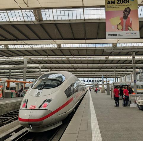 Ein weißer ICE-Hochgeschwindigkeitszug der Deutschen Bahn mit charakteristischem roten Streifen steht frontal am Bahnsteig einer überdachten Bahnhofshalle (Kopfbahnhof). Rechts auf dem gepflasterten Bahnsteig stehen Bahnmitarbeiter in roter Arbeitskleidung neben einem kleinen Kiosk. Von der Decke der weitläufigen Stahl-Glas-Konstruktion hängt ein großes Werbeplakat für den Bezahldienst „Wero“ mit der Aufschrift „AM ZUG!“. Im Hintergrund sind weitere Gleise, Züge und eine Glasfassade mit einem G…