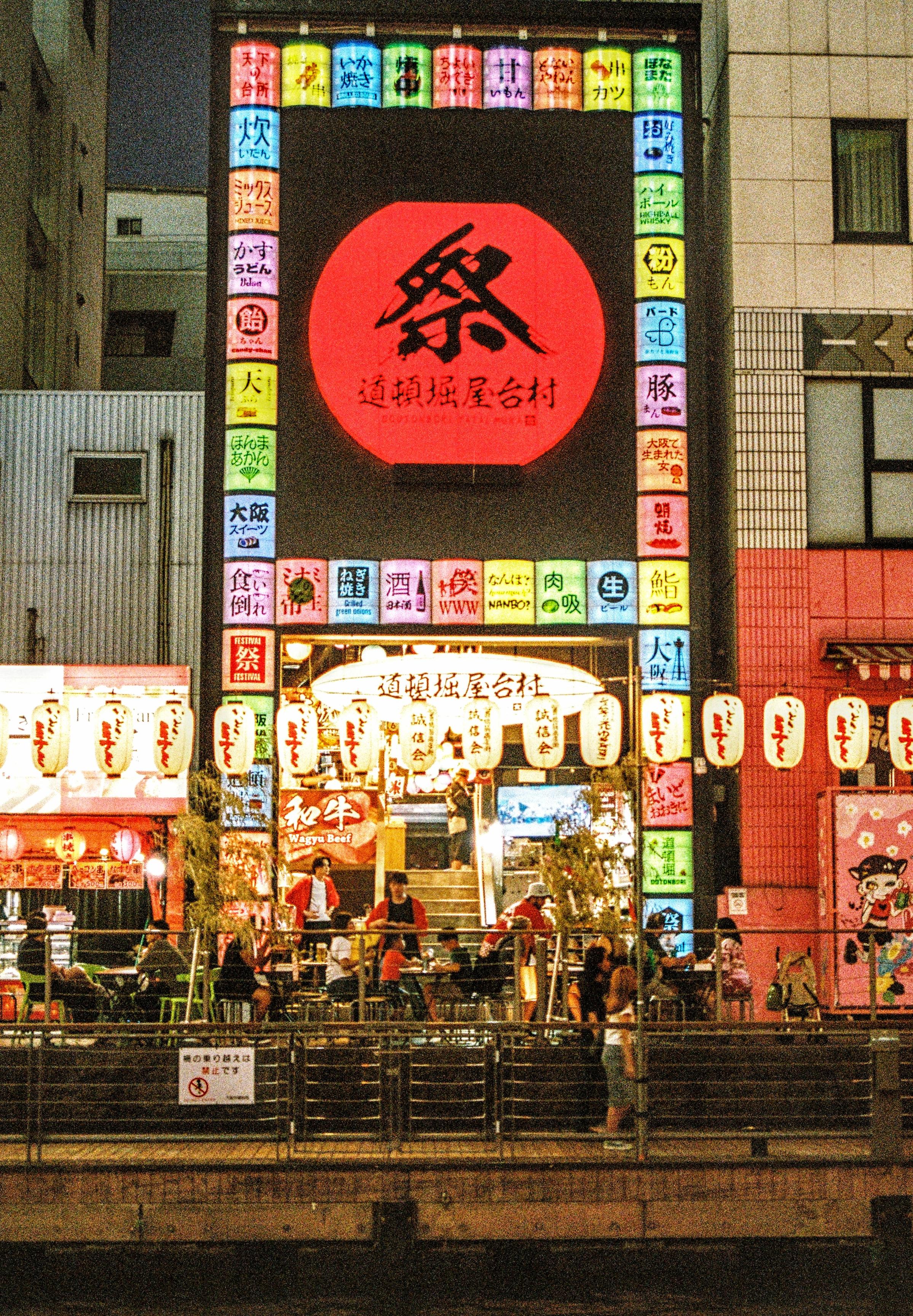 Colour 35mm film photograph of a restaurant by the Dotonbori Canal in Osaka, Japan. Image shows a brightly lit multi coloured entrance with people enjoying their meals. A young lady is walking along the footpath on the canal below. Taken July 2025.