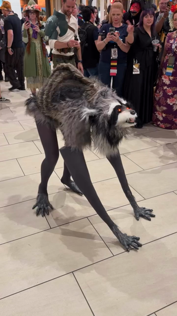 In a convention hall, a person in costume as the Internet meme and raccoon Jimothy, complete with raccoon compact body, face, long arms and legs (the person’s).