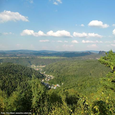 Weitläufige Hügellandschaft mit dichten Wäldern und einem kleinen Ort im Tal, dessen Häuser sich entlang einer Straße durch das Grün ziehen. Am Horizont wechseln sich bewaldete Erhebungen mit offenen Feldern ab. Rechts in der Ferne ragen markante Felsformationen über die Baumwipfel. Der Himmel ist blau mit einigen verstreuten weißen Wolken.