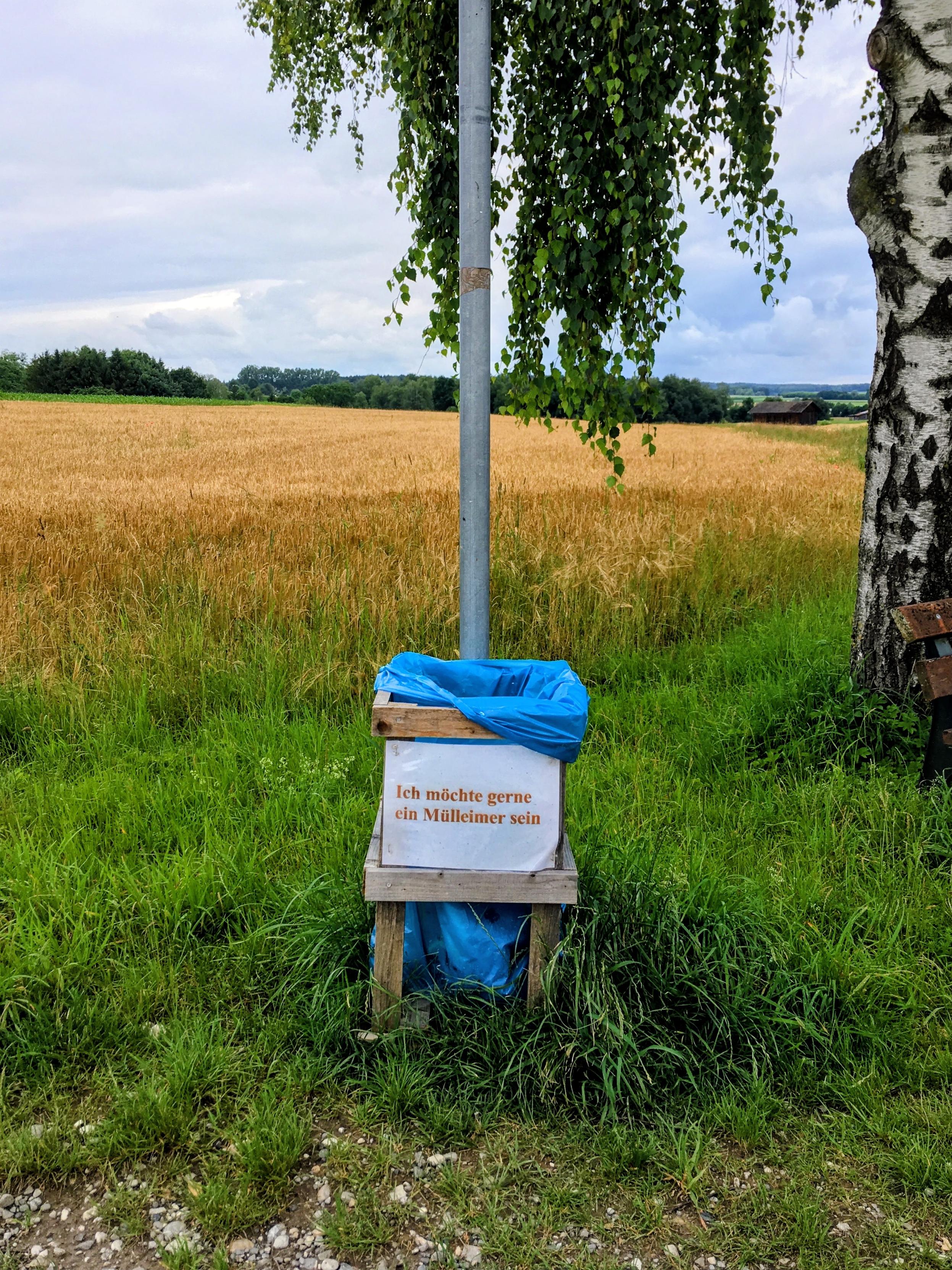 Selbstgebauter Mülleimer aus Holzlatten mit blauem Müllbeutel an einem Metallpfosten am Feldrand. Darauf ein Schild mit der Aufschrift: „Ich möchte gerne ein Mülleimer sein“.