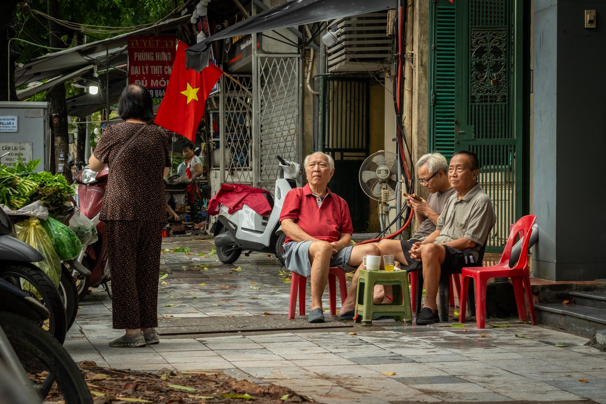 Three elderly gentlemen sitting in the street on small plastic chairs look at a lady standing a few meters away.