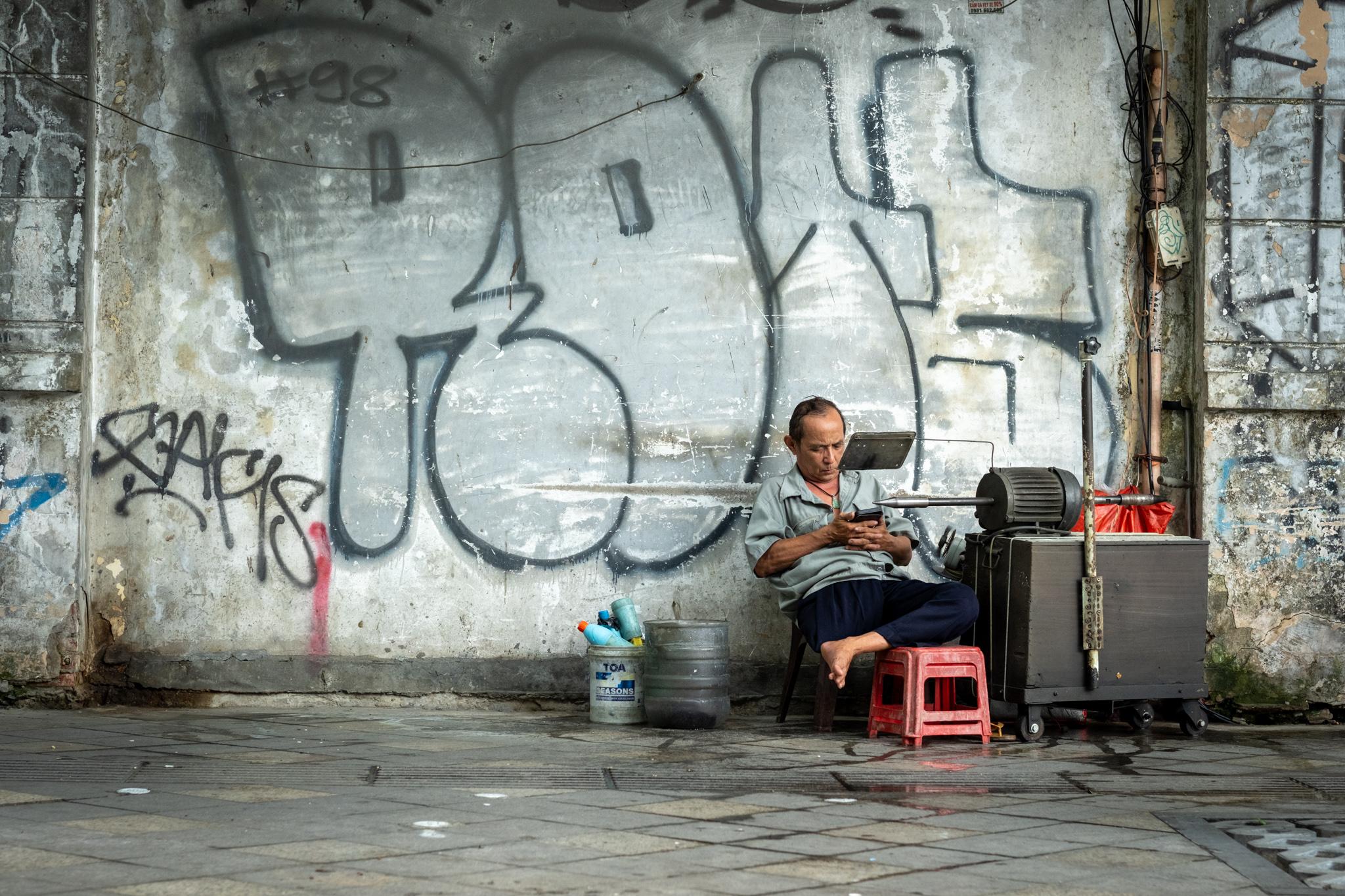 A man waits for customers. He is seated next to a machine that looks like a lathe of some kind. There's graffiti on the concrete wall behind him.