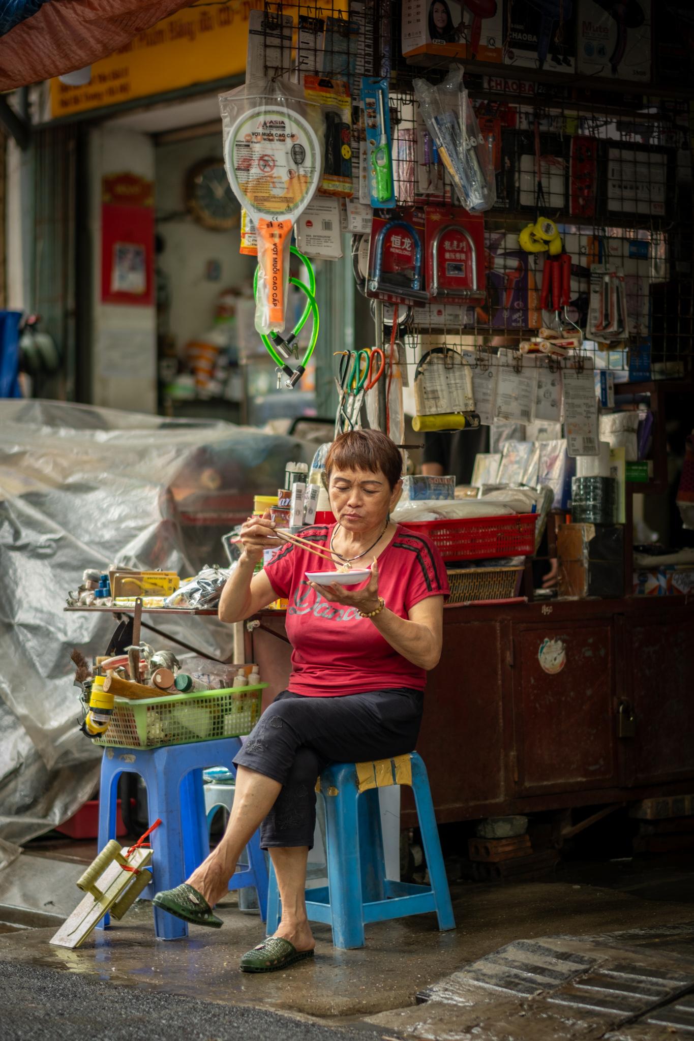 A shopkeeper eats breakfast in front of her little shop that appears to sell various household items and tools.
