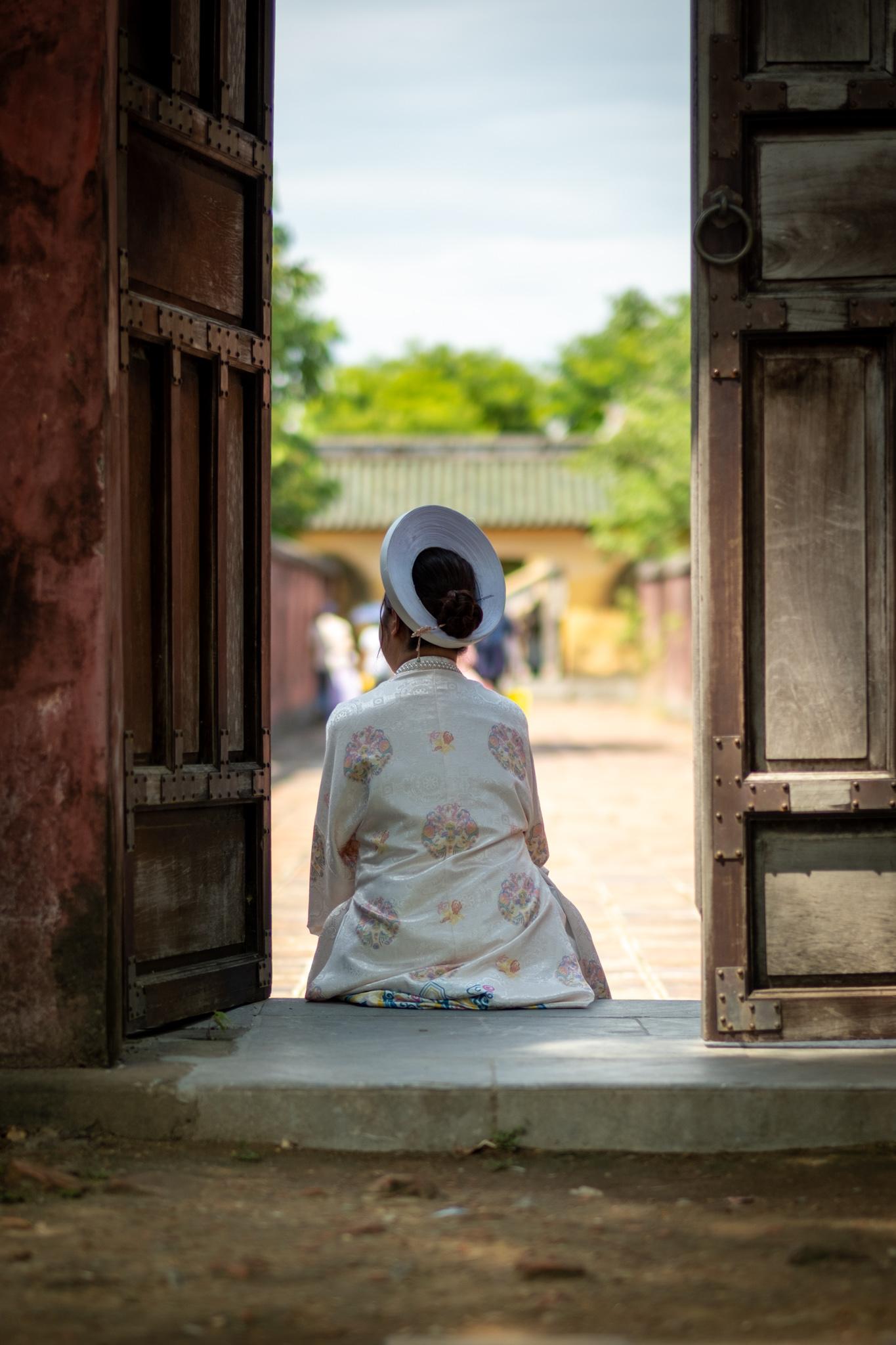 A woman in traditional clothing rests in a doorway during September 2nd celebrations in the Citadel in Hue. We see her from the back. Her clothing is mostly white.