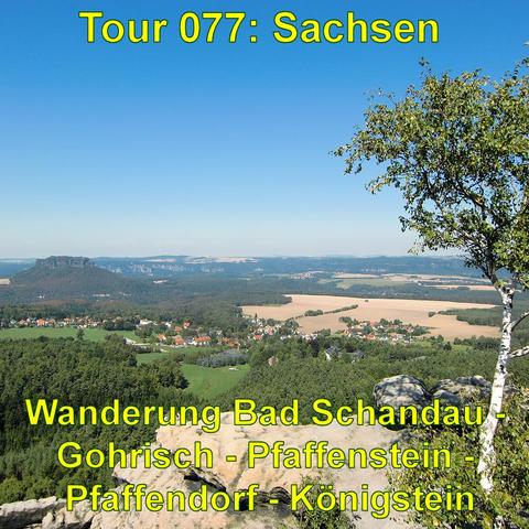 Das Bild bietet einen weiten Blick über eine hügelige Landschaft. Im Vordergrund ist der Rand eines Felsplateaus zu sehen, das dicht mit Bäumen bewachsen ist, darunter auch ein hervorstechender Baum mit grünen Blättern auf der rechten Seite. In der Ferne erstrecken sich Felder und Wälder, unterbrochen von einem kleinen Dorf mit zahlreichen Häusern. Im Hintergrund erheben sich weitere bewaldete Hügel, und der blaue Himmel ist weitgehend klar mit nur wenigen Wolken. Die Szenerie wirkt ruhig und m…