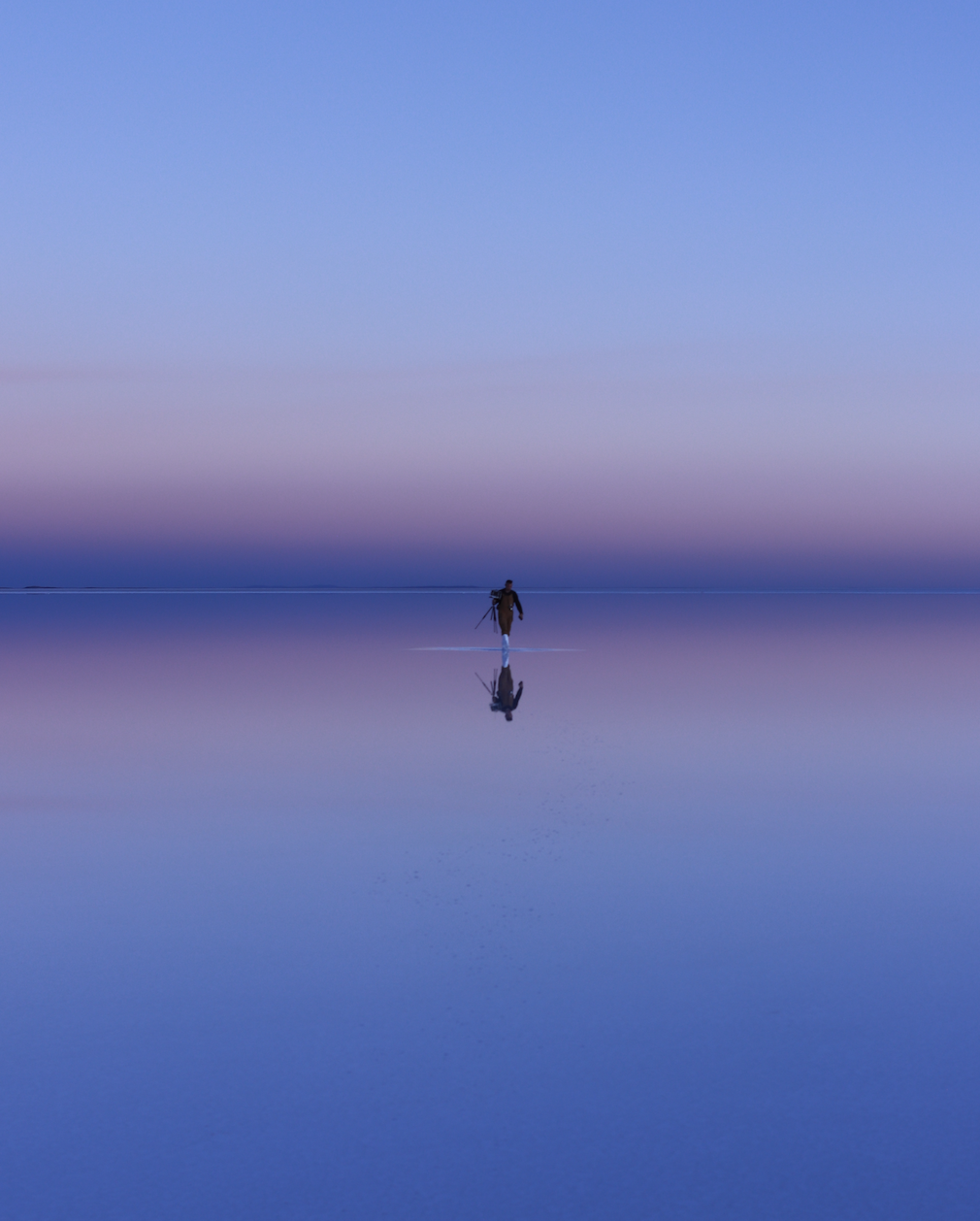 a series of horizontal colors fade into each other, staring with light blue to a peachy yellow and thin pinks, followed by a deep purple. this is mirrored on the bottom half of the image. a small figure walks in the center, far in the distance