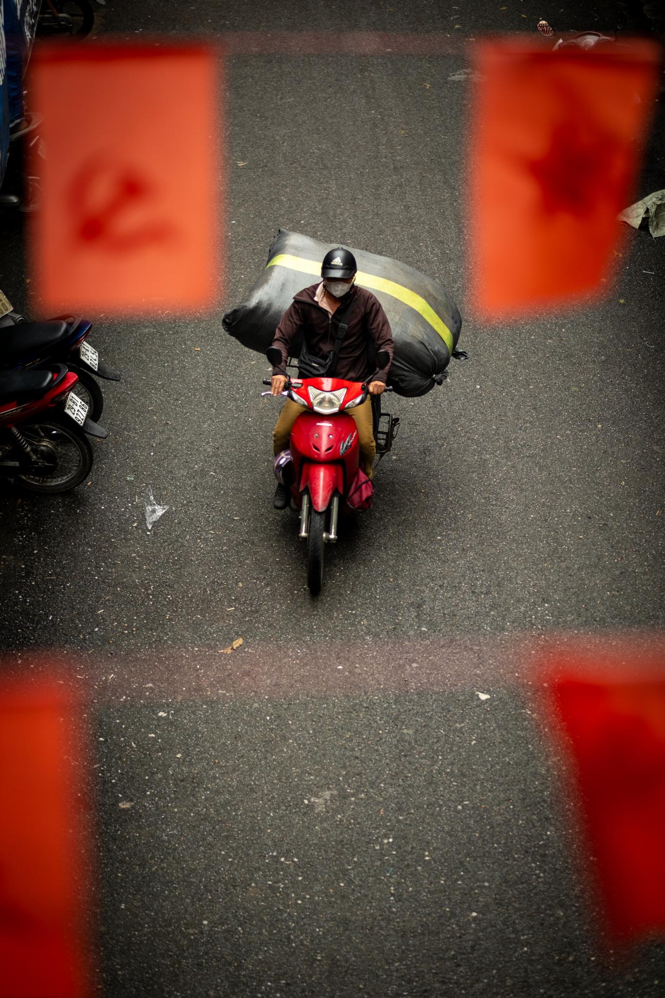 A delivery driver, seen from above, rides a red scooter below lines with small red flags. Stars and hammer/sickle symbols can be seen on on the flags.