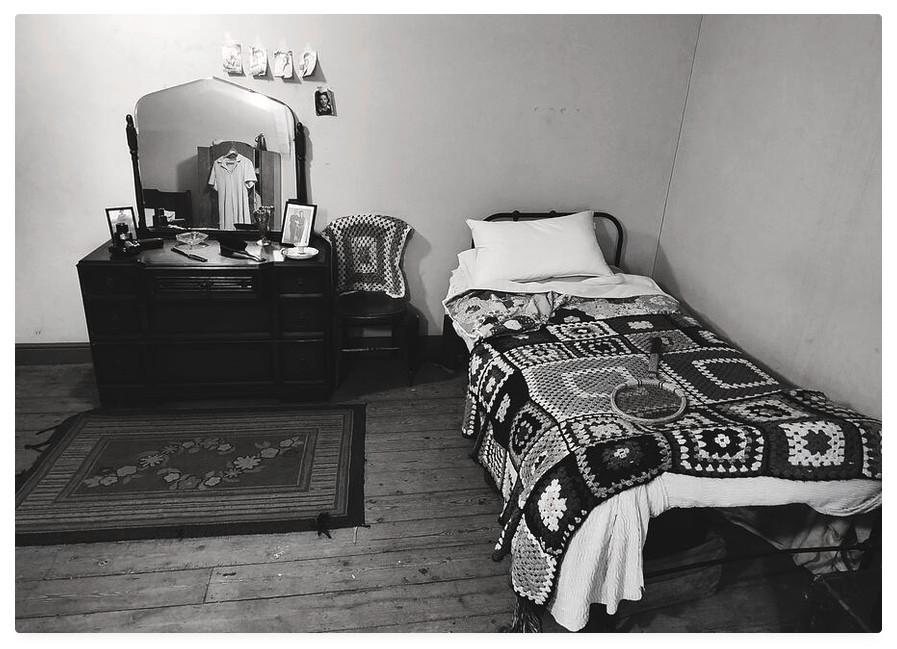 Vintage looking black and white photograph of a bedroom from the early mid 20th century; with a crochet blanket covered single bed and a wooden dressing table.