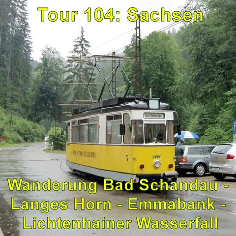 Eine gelbe Straßenbahn mit der Aufschrift Bad Schandau und Kirnitzschtalbahn steht auf Schienen in einer von Bäumen gesäumten Straße. Es regnet, und ein Fußgänger mit einem blauen Regenschirm ist im Hintergrund sichtbar. Neben der Straßenbahn sind geparkte Autos zu erkennen.