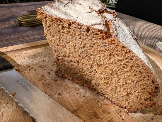 Nahaufnahme eines frisch angeschnittenen Laibs dunklen Bauern- oder Vollkornbrots. Die Schnittfläche zeigt eine dichte, gleichmäßig geporte Krume in warmem Braun. Die Oberseite des Brotes hat eine leicht rissige, kräftig ausgebackene Kruste, die mit weißem Mehl bestäubt ist. Das Brot steht schräg auf einem Holzschneidebrett mit Rillen, auf dem feine Mehlspuren und Krümel liegen. Im linken Vordergrund liegt die gezackte Klinge eines Edelstahl-Brotmessers mit schwarzem Griff. Der Hintergrund zeig…
