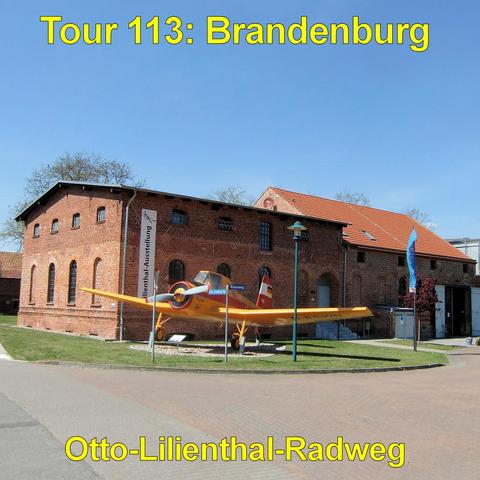 Rotes Backsteingebäude mit Satteldach und mehreren rundbogigen Fenstern. Vor dem Gebäude steht auf einer kleinen Grünfläche ein gelb lackiertes Propellerflugzeug mit starrem Fahrwerk und niedrigen Tragflächen. Das Flugzeug ist als Ausstellungsstück aufgestellt und von kleinen Schildern umgeben. Neben dem Gebäude befinden sich Fahnenmasten und eine Eingangstür mit Treppe. Ein gepflasterter Weg verläuft im Vordergrund entlang der Anlage. Der Himmel ist klar und blau, Bäume stehen hinter dem Gebäu…
