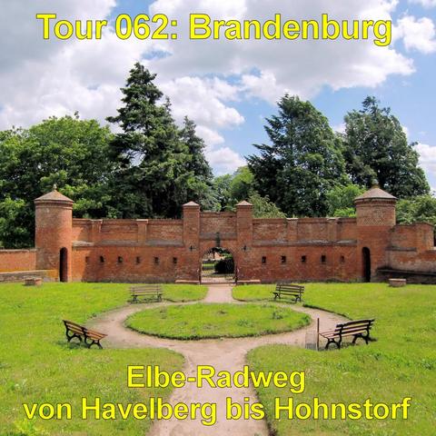 Eine niedrige, historische Backsteinmauer mit zwei runden Türmen und kleinen spitzen Dächern steht in einer grünen Parkanlage. In der Mitte befindet sich ein rundbogiges Tor mit einem geöffneten Gitter, durch das ein Weg hindurchführt. Die Mauer ist gleichmäßig gemauert und von kleinen rechteckigen Öffnungen durchzogen. Davor liegt eine Rasenfläche mit einem kreisförmig angelegten Weg. Mehrere Holzbänke stehen symmetrisch entlang des Rundwegs. Hohe Laub- und Nadelbäume ragen hinter der Mauer em…
