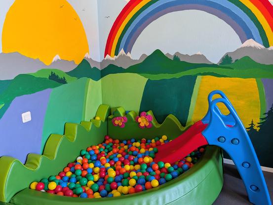 A ball pit for children in a corner of the room, with a small slide in front of a colorfully painted wall depicting mountains, meadows, the sun, and a rainbow.