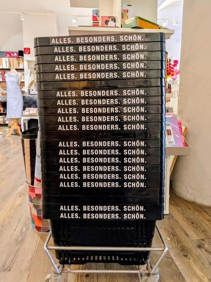 In einem hell beleuchteten, modernen Einrichtungsgeschäft steht ein hoher Stapel übereinander gestapelter, schwarzer Plastikkisten auf einem Metalltrolley mit Rollen auf einem hellen Holzboden. Auf der Vorderseite jeder Kiste ist der Schriftzug "ALLES. BESONDERS. SCHÖN." in weißer, sauberer, serifenloser Schrift aufgedruckt. Der Stapel dominiert die Mitte des Bildes. Im Hintergrund links ist eine Schaufensterpuppe mit einer grauen Schürze und ein Regal mit verschiedenen Dekoartikeln und Büchern…